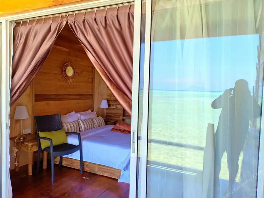 An exterior view of a bedroom at Dreamy Wave Dwelling Resort showing a bed facing seamless glass-panel doors, authentic wooden walls with glossy hardwood floors, and air con.