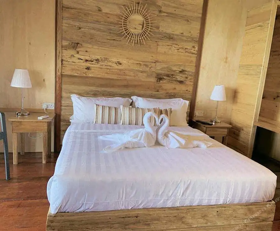 A rustic bedroom entirely clad in natural wood, featuring a cozy bed with towel swans, matching bedside tables with lamps, and a decorative sunburst mirror, exemplifying traditional timber architecture at Dreamy Wave Dwelling Resort.